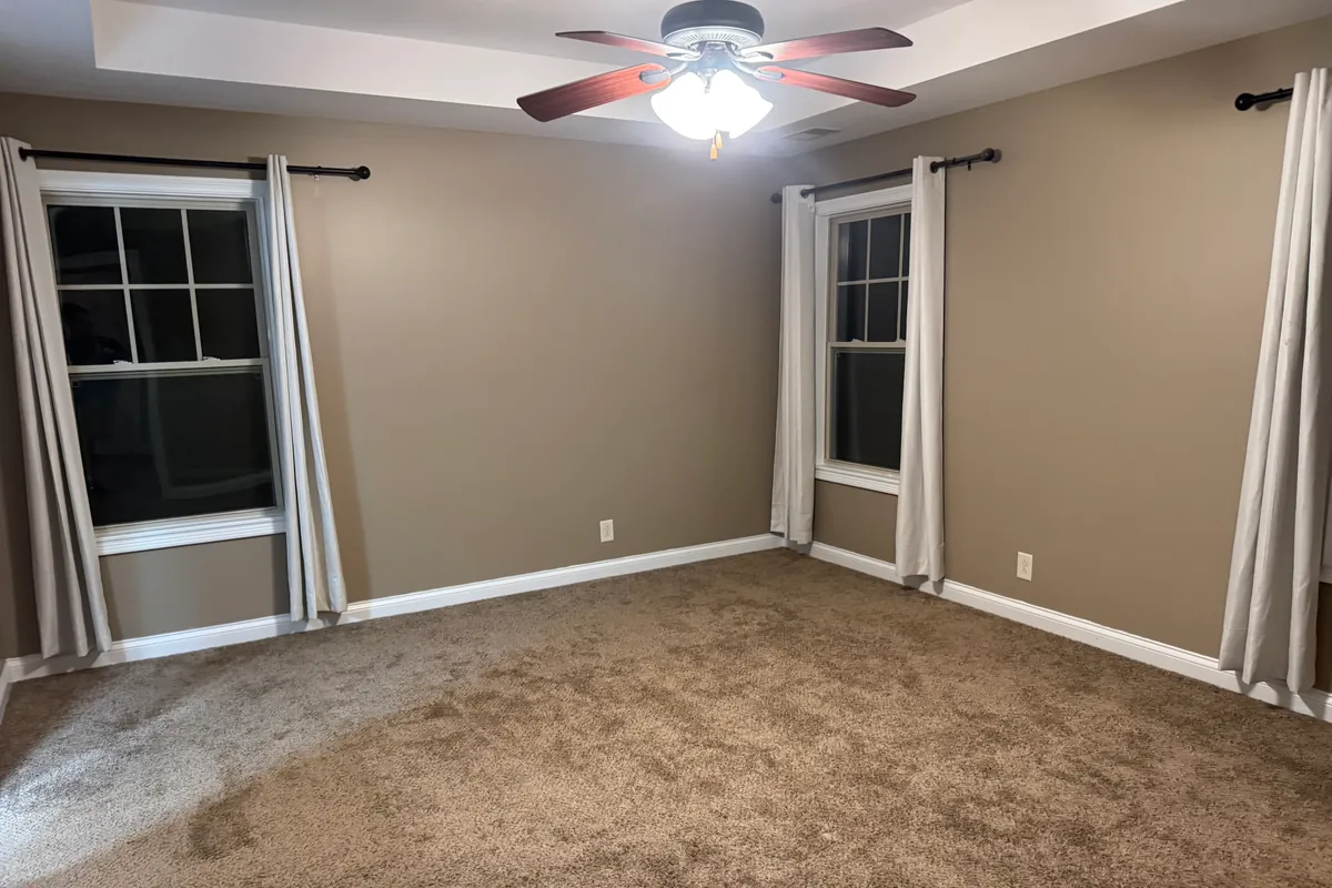 Empty room cleared down to bare carpet, with tan walls, white trim, a tray ceiling, a ceiling fan and two curtained windows. Nothing left in the room.